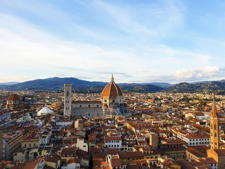 Vista aérea de una ciudad histórica, centrándose en una catedral con cúpula roja y campanario, con montañas visibles en el horizonte.