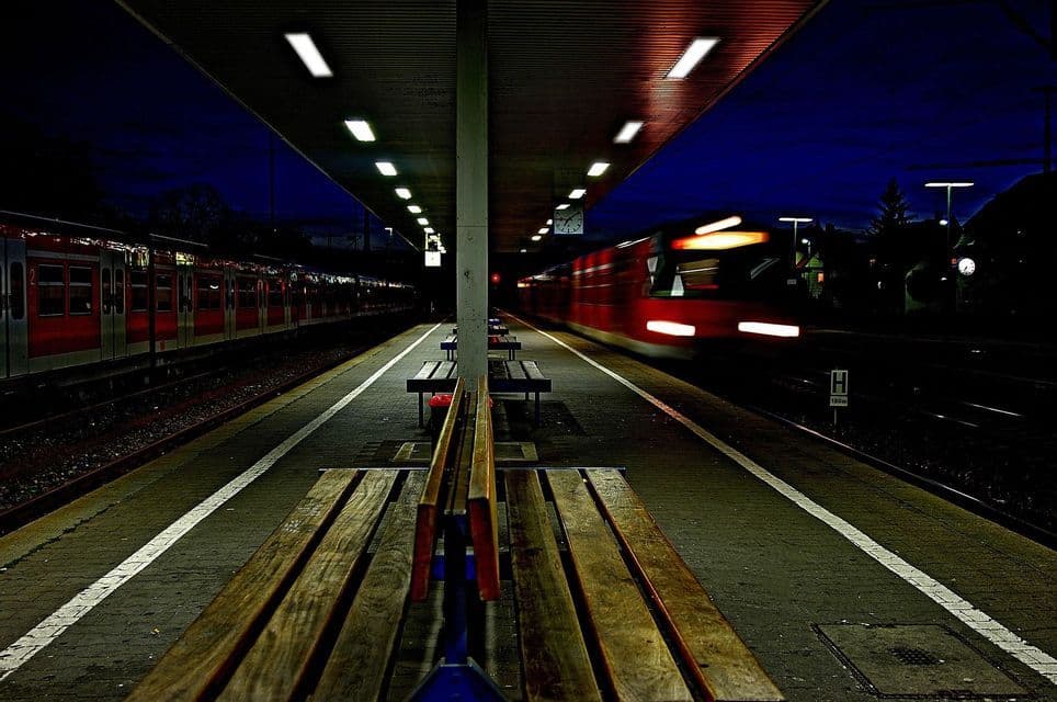 Un treno rosso crea un effetto mosso mentre sfreccia di notte davanti a una banchina della stazione vuota e illuminata, con un altro treno fermo di fronte.