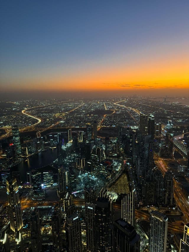 Veduta aerea di una metropoli estesa al crepuscolo, con grattacieli e autostrade illuminati sotto un cielo sfumato arancione e blu.