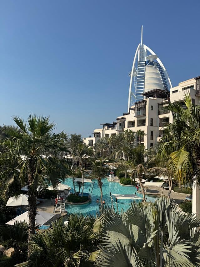 Vista di una piscina di resort con palme, con l'hotel Burj Al Arab visibile sullo sfondo contro un cielo azzurro e limpido.