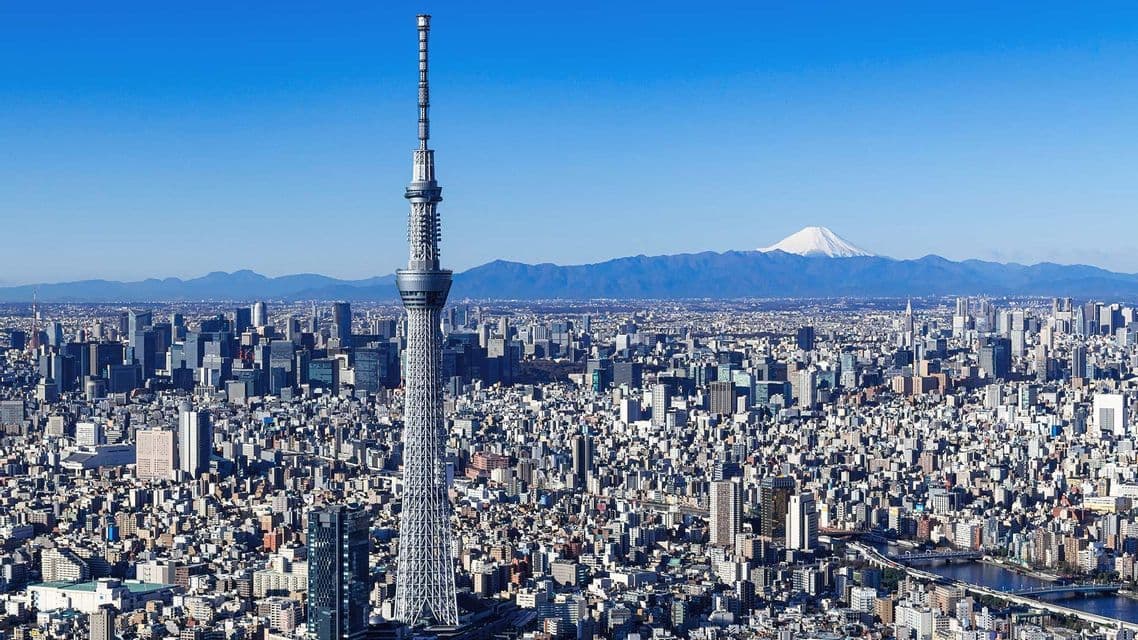 Un'alta torre delle comunicazioni domina un denso paesaggio urbano, con una montagna innevata visibile in lontananza.
