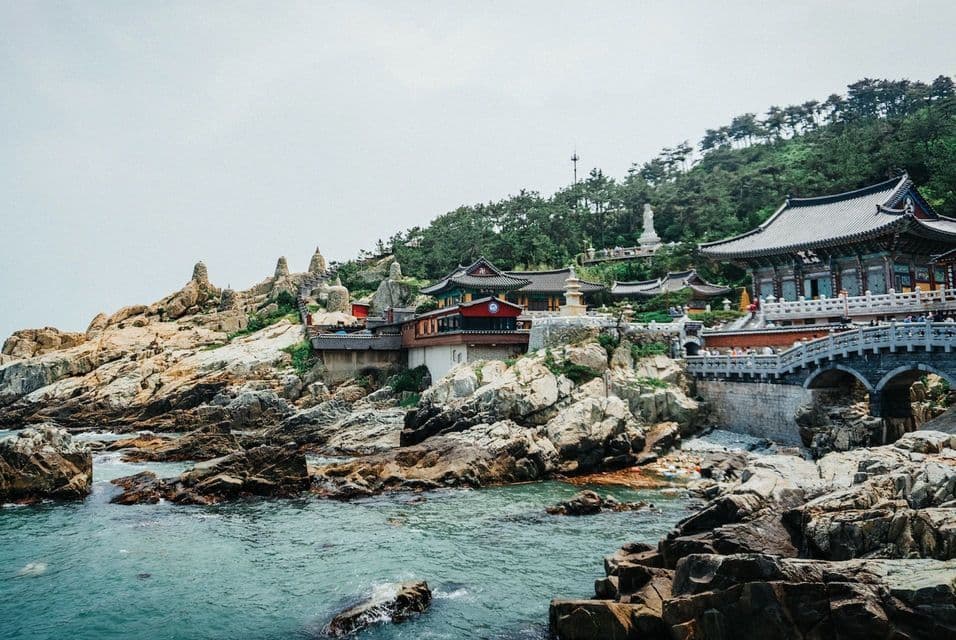 Un tradizionale complesso di templi con un ponte di pietra, edificato su una costa rocciosa vicino ad acque turchesi, sotto un cielo coperto.