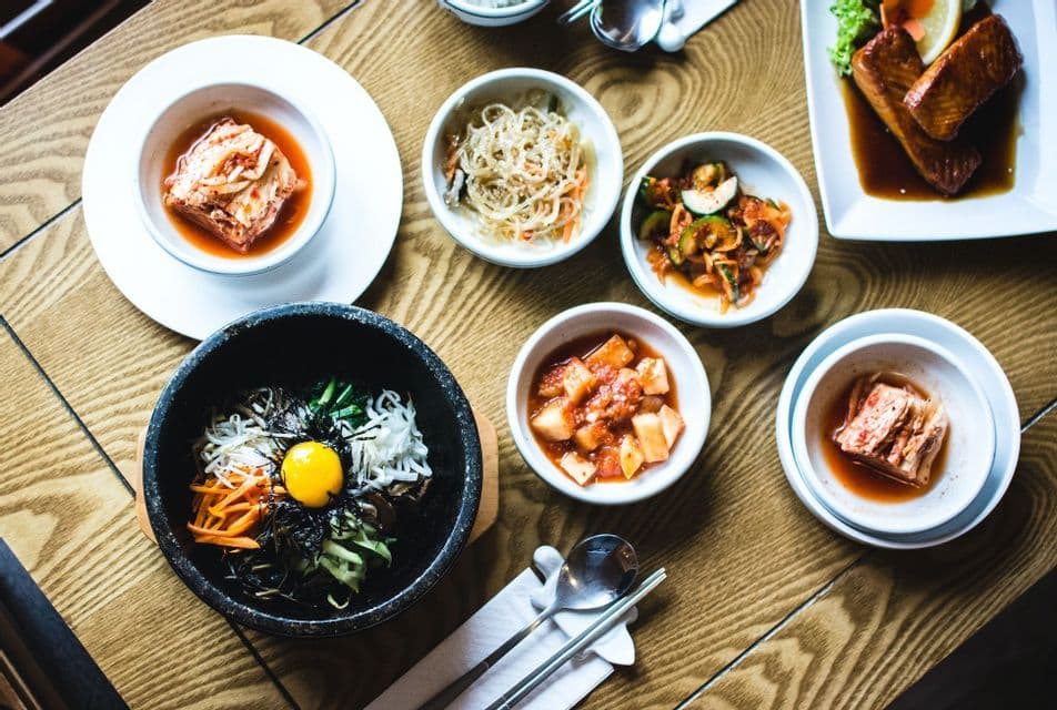 Una vista dall'alto di un tradizionale pasto coreano, che include bibimbap con un tuorlo d'uovo e vari contorni, il tutto disposto su un tavolo di legno.