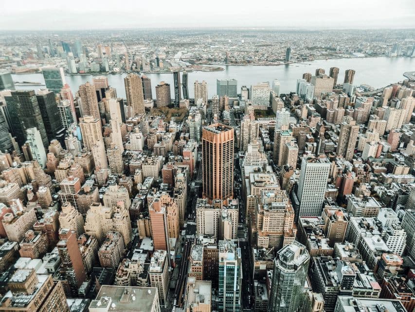 Luftaufnahme einer dichten Stadtlandschaft mit Wolkenkratzern und einem breiten Fluss im Hintergrund an einem bewölkten Tag.