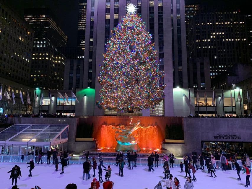 Der Weihnachtsbaum des Rockefeller Centers, hell erleuchtet mit bunten Lichtern, überragt nachts die Menschen, die auf der Eisbahn darunter Schlittschuh laufen.