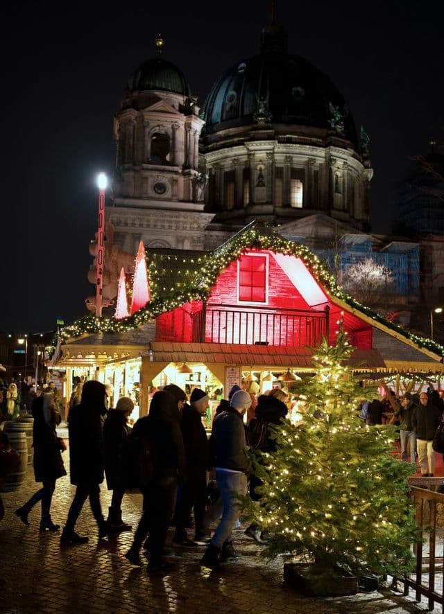 Menschen gehen nachts über einen Weihnachtsmarkt, vorbei an beleuchteten Ständen und einem beleuchteten Baum, mit einem großen Kuppelgebäude im Hintergrund.