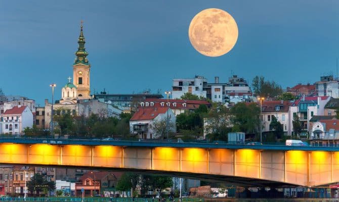 Ein großer, goldener Vollmond hängt am Dämmerungshimmel über einer Stadtlandschaft, mit einer beleuchteten Brücke im Vordergrund und einem dahinter aufragenden Kirchturm.