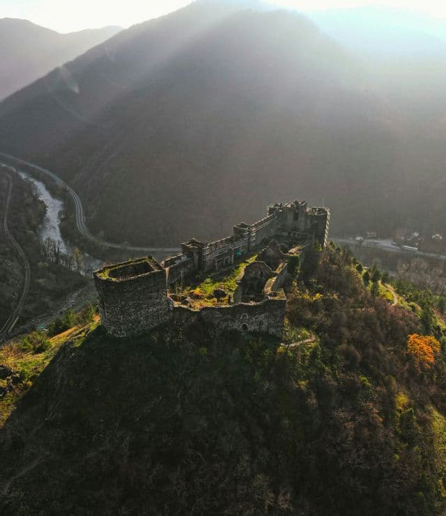Una vista aerea delle rovine di un castello in pietra su una ripida collina boscosa che si affaccia su una valle fluviale, con il sole che spunta sulle montagne lontane.