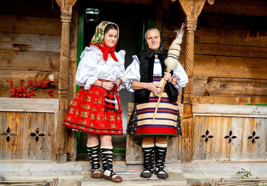 Deux femmes en costumes folkloriques traditionnels se tiennent sur un porche en bois, l'une tenant une quenouille pour filer la laine.