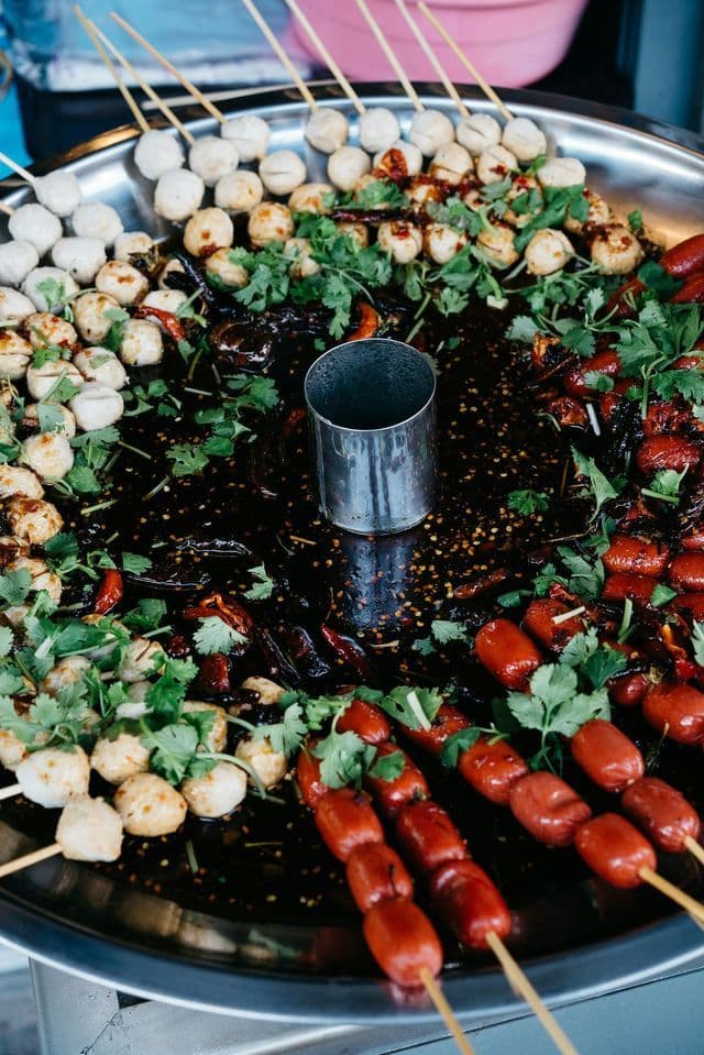 Brochettes de boulettes de viande et de petites saucisses, présentées sur un plateau métallique, nappées d'une sauce sombre et épicée et garnies de coriandre.