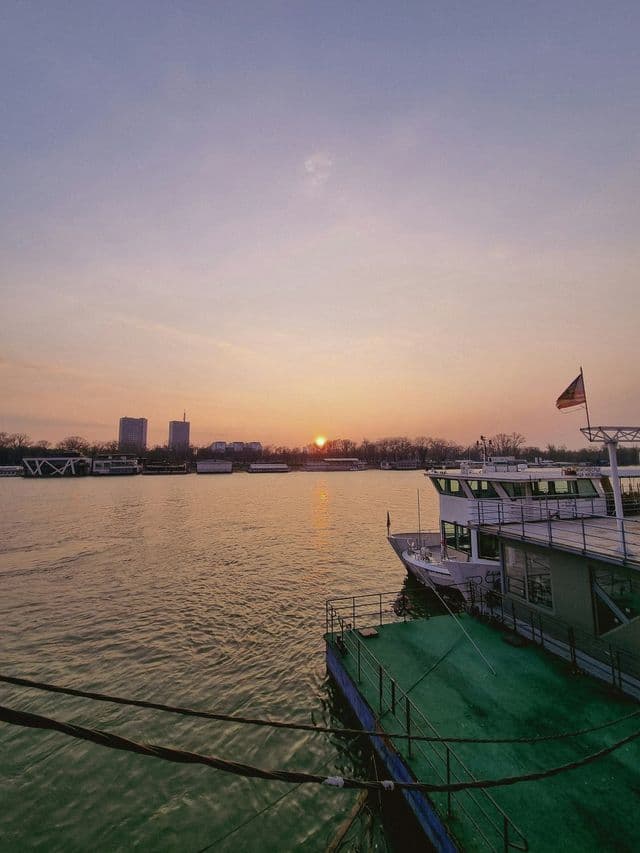 Le soleil se couche sur une large rivière avec des bateaux amarrés le long de la berge et une ligne d'horizon urbaine visible au loin.