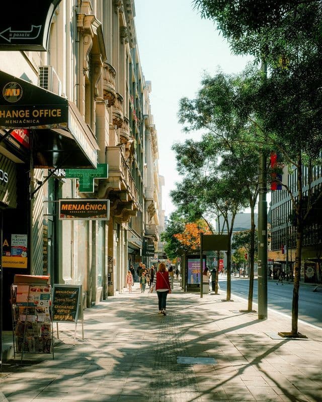 Une femme vêtue d'un gilet rouge marche sur un trottoir ensoleillé, bordé d'immeubles ouvragés et d'arbres verdoyants.