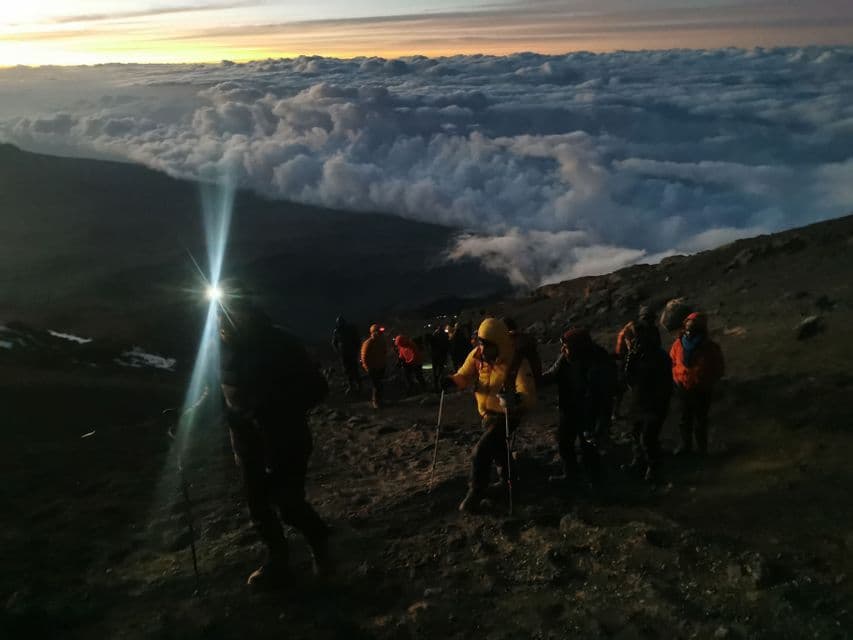 Un viaggio di gruppo WeRoad in trekking su un sentiero di montagna roccioso con lampade frontali all'alba, in alto sopra un mare di nuvole.