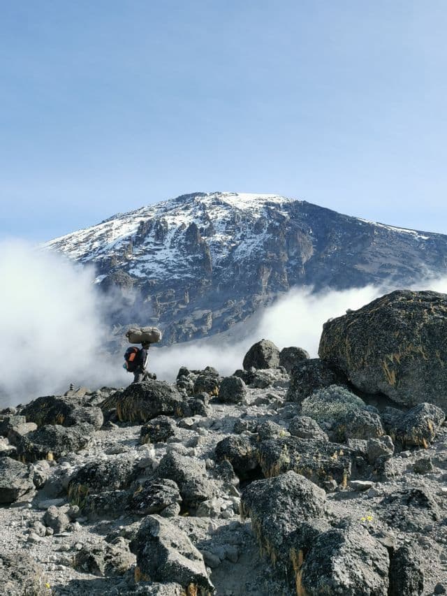 Un portatore con un grande fardello sulla testa e uno zaino si erge su un sentiero roccioso con una cima montuosa innevata che svetta tra le nuvole.
