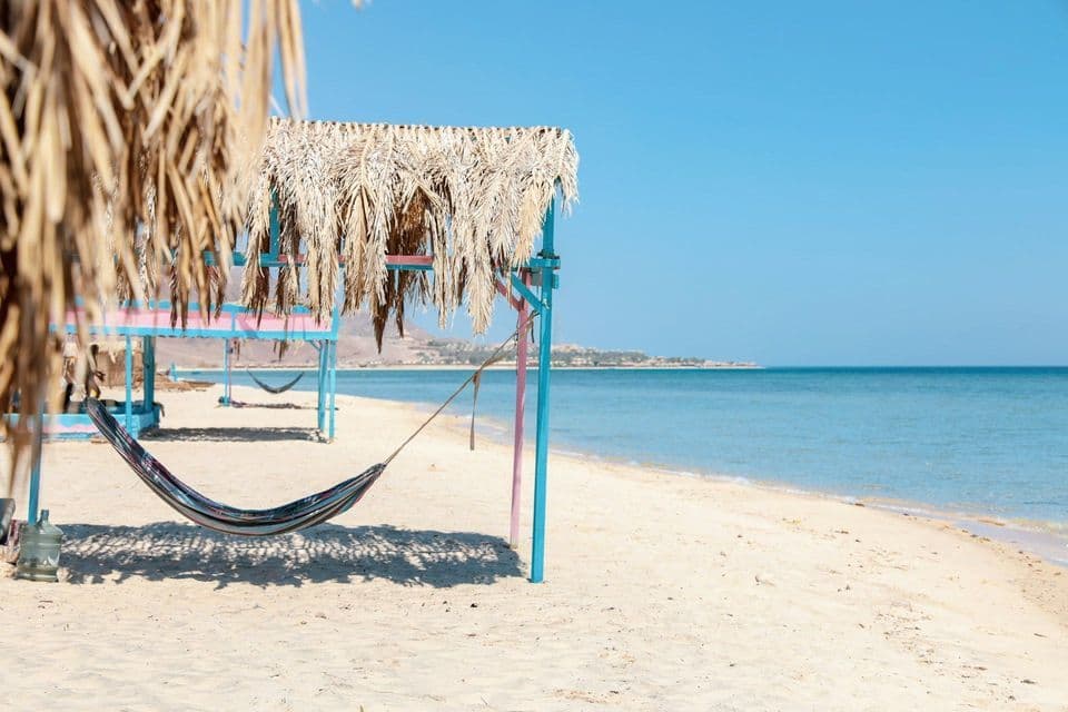 An empty hammock hangs from a colorful structure with a thatched roof on a sandy beach next to the calm sea.