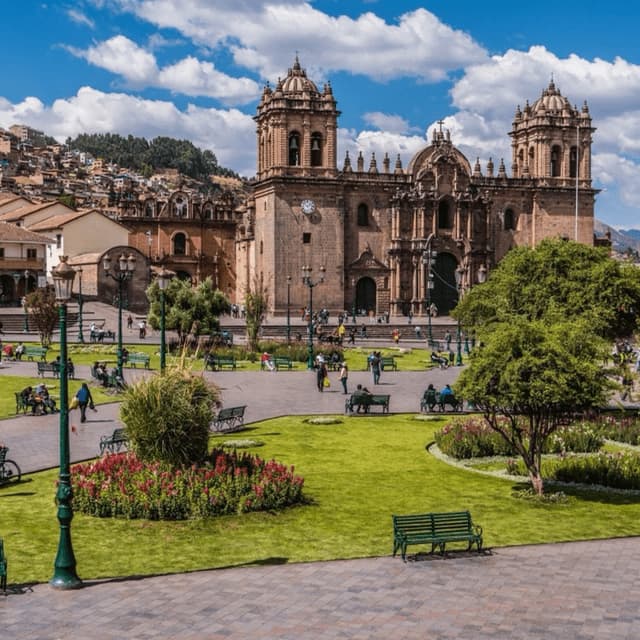 A large stone cathedral with two bell towers stands in a sunny public square with green grass, gardens, and people relaxing.