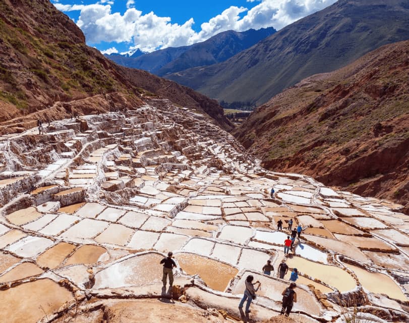 A WeRoad group trip walks among terraced salt pans built into a mountain valley under a bright blue sky.