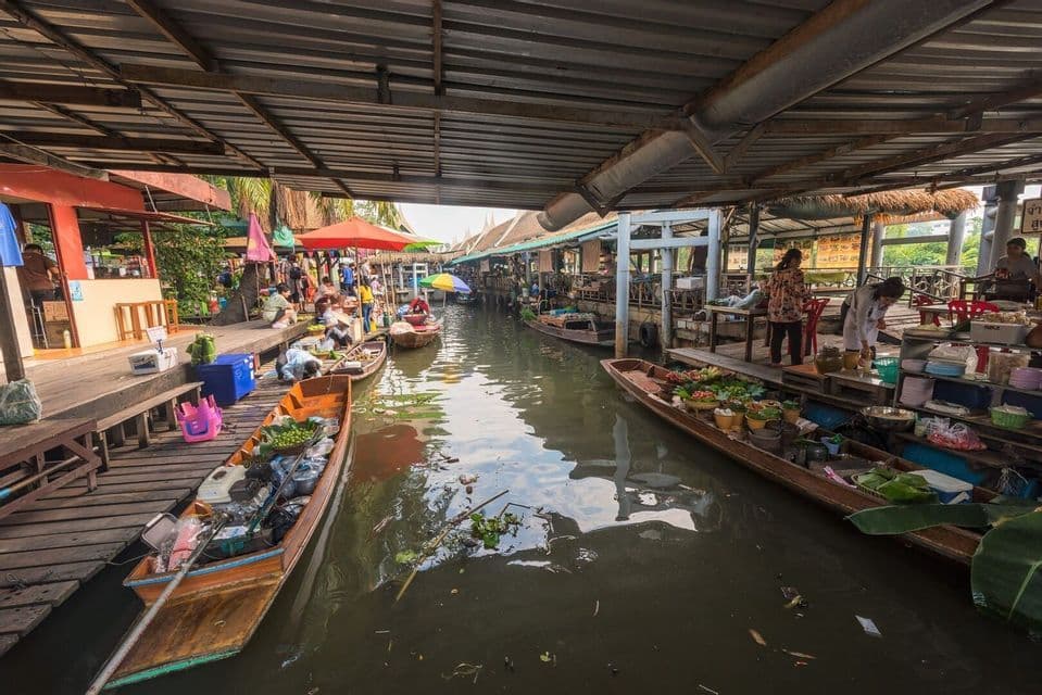 Largas barcas de madera con vendedores y productos flotan en un canal de un animado mercado cubierto.