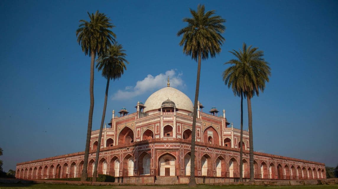 Un grande edificio simmetrico in pietra, rosso e bianco, con una cupola centrale, è incorniciato da quattro alte palme sotto un cielo azzurro.
