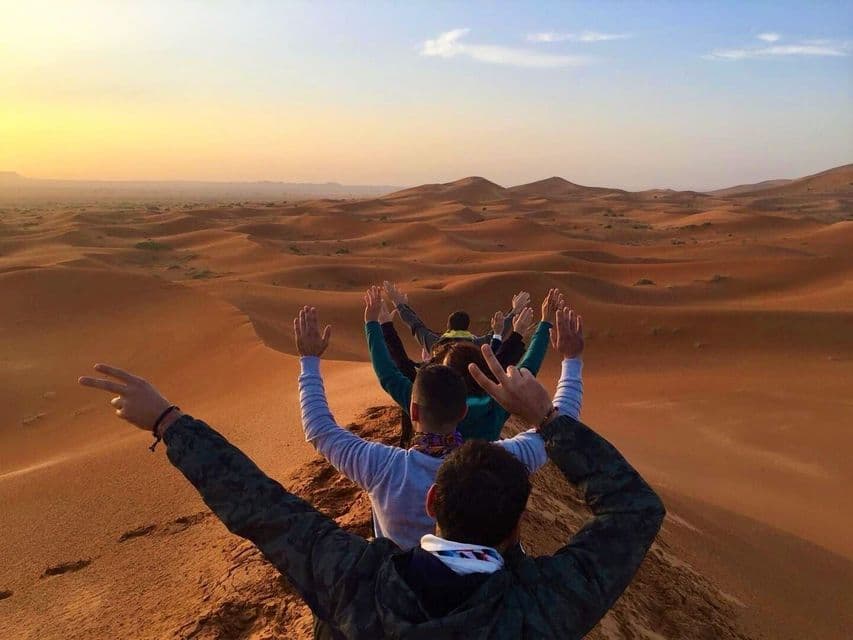 Un gruppo WeRoad, visto di spalle, siede su una duna di sabbia con le braccia alzate, ammirando un vasto deserto al tramonto.