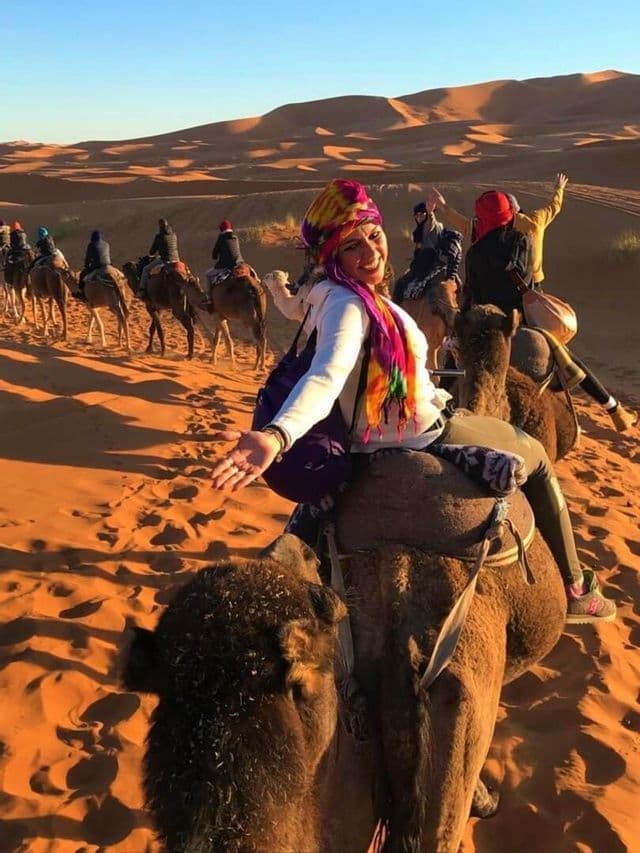 Una mujer en camello sonríe a la cámara mientras su grupo de viaje WeRoad la sigue en fila a través de las dunas de arena del desierto.