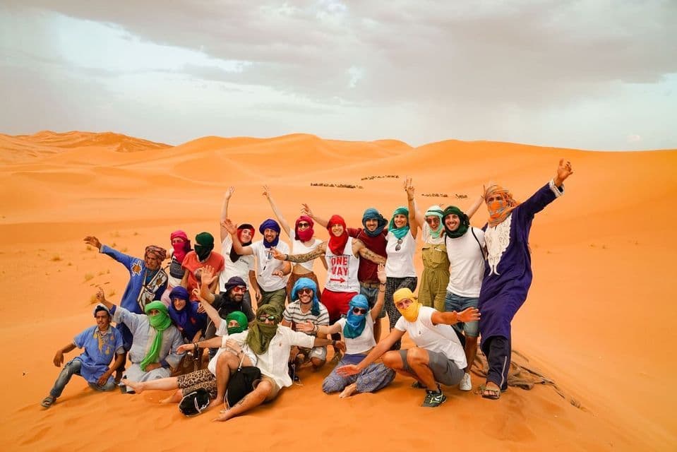 Un viaje en grupo de WeRoad de personas con pañuelos de colores posa para una foto en dunas de arena naranja del desierto.