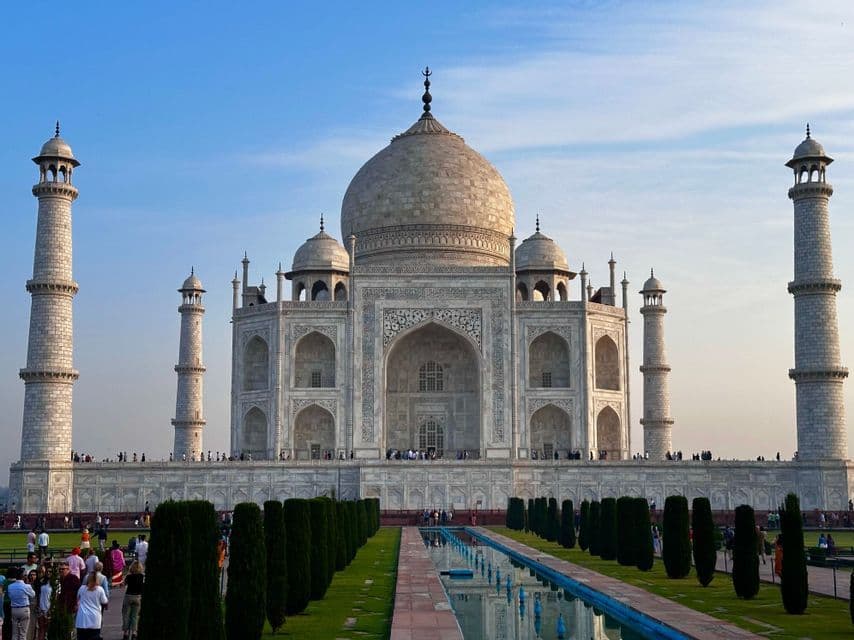 Der Taj Mahal steht hinter seinem Spiegelbecken und den Gärten, während Touristen unter einem klaren blauen Himmel über das Gelände spazieren.