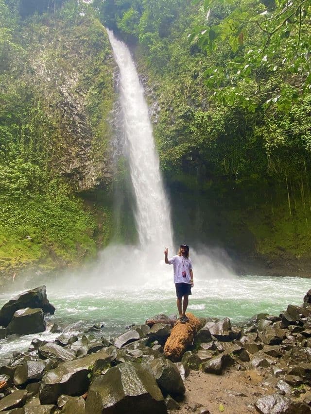 Une personne aux cheveux longs se tient sur un rocher, faisant un signe de paix devant une haute cascade dans une jungle luxuriante et verte.