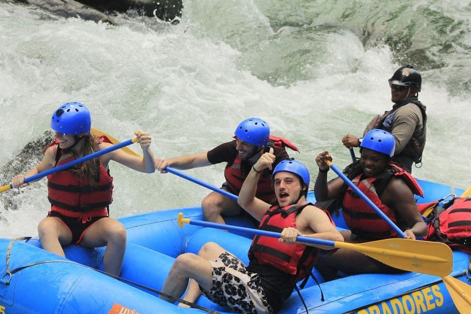Un grupo de WeRoad sonríe mientras hace rafting en rápidos de aguas bravas en un bote azul, usando cascos y chalecos salvavidas.
