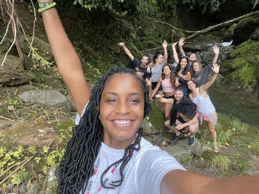 Una mujer sonriente con rastas se toma una selfie con su grupo de viaje de WeRoad mientras posan junto a un arroyo rocoso en un bosque exuberante.