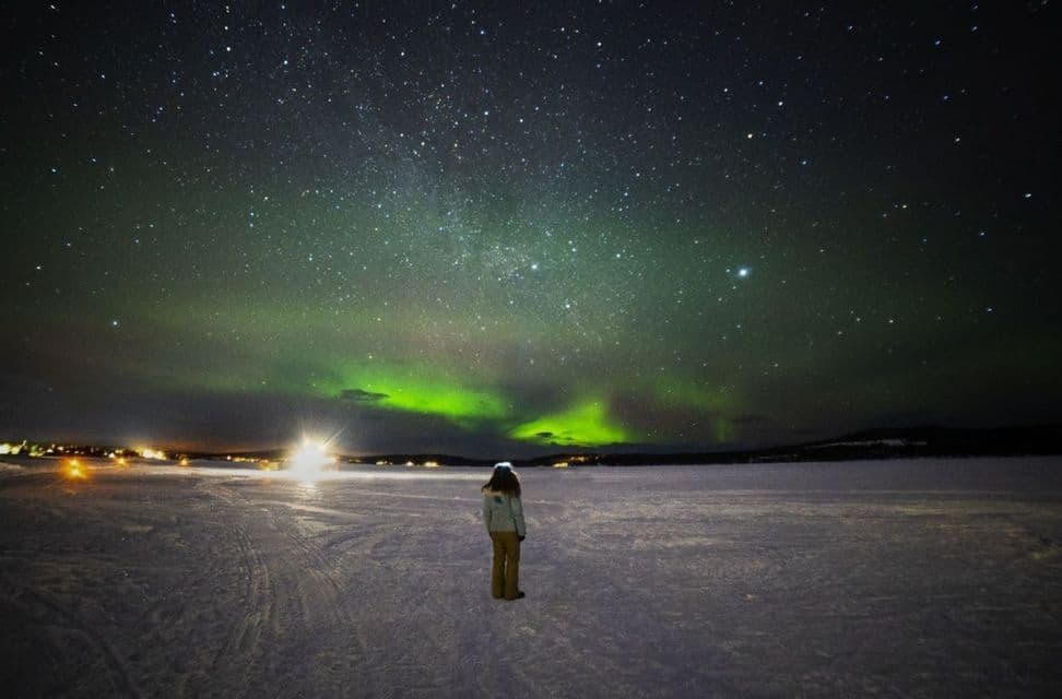 Eine Person steht nachts in einer verschneiten Landschaft und blickt zu den grünen Nordlichtern und einem Sternenhimmel auf.