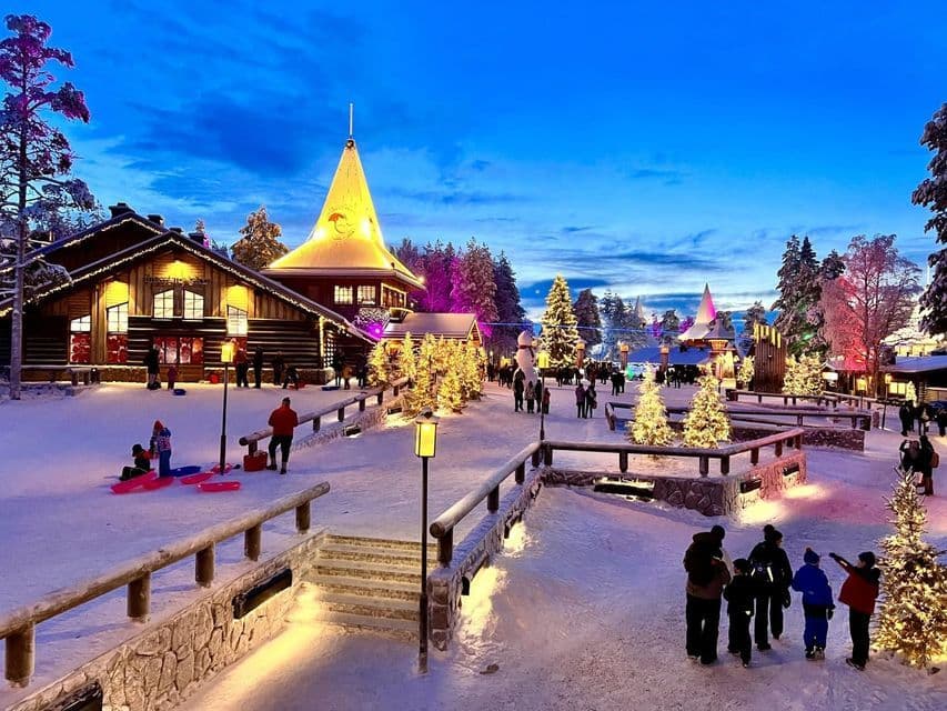 A snowy village at dusk with people walking among illuminated log cabins and decorated Christmas trees.