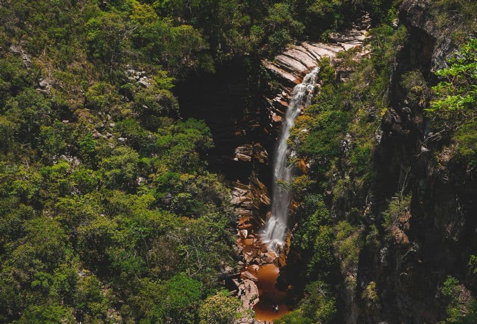 Una vista dall'alto di una cascata che si getta da una scogliera rocciosa in un laghetto rossastro, circondata da una lussureggiante foresta verde con persone radunate.