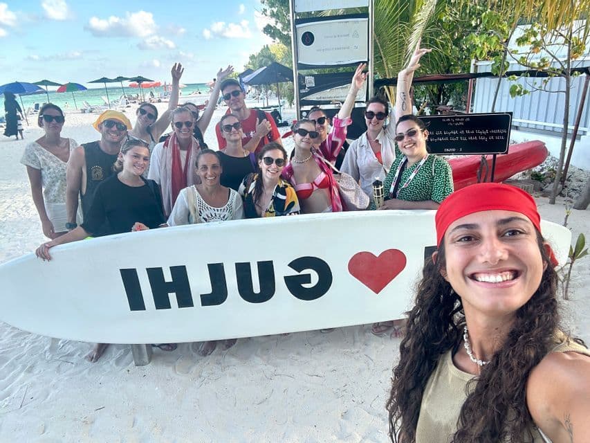 Un voyage de groupe WeRoad pose pour un selfie souriant sur une plage ensoleillée, tenant une grande planche de surf blanche.
