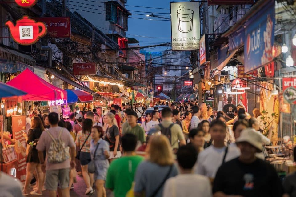 A large crowd of people walks through a narrow, bustling street market at dusk, surrounded by illuminated signs and food stalls.