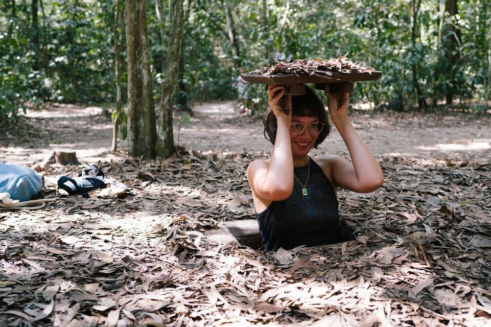 A smiling woman with glasses emerges from a leaf-covered hole in the ground in a forest, holding the lid above her head.