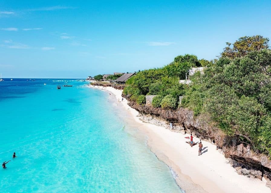 Una vista aerea di una spiaggia tropicale con sabbia bianca, acqua turchese e una scogliera verde lussureggiante sotto un cielo azzurro limpido.