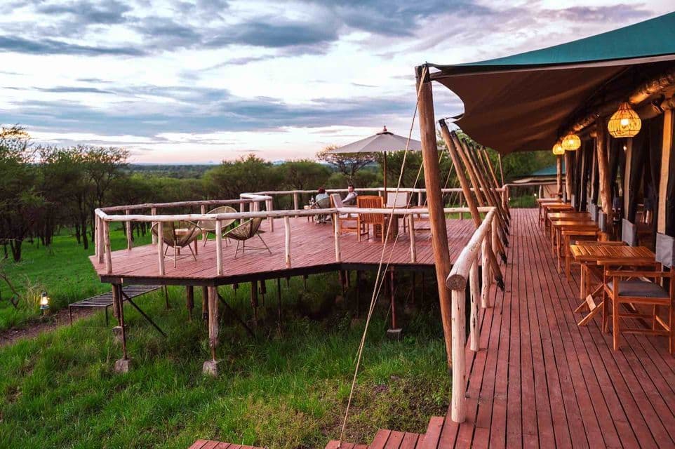 Una terrazza rialzata in legno di un safari lodge, con tavoli e sedie, che si affaccia su un paesaggio erboso e alberato sotto un cielo nuvoloso.