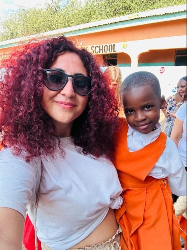 Una donna con capelli rossi ricci e occhiali da sole scatta un selfie con un bambino piccolo in uniforme scolastica davanti a una scuola.