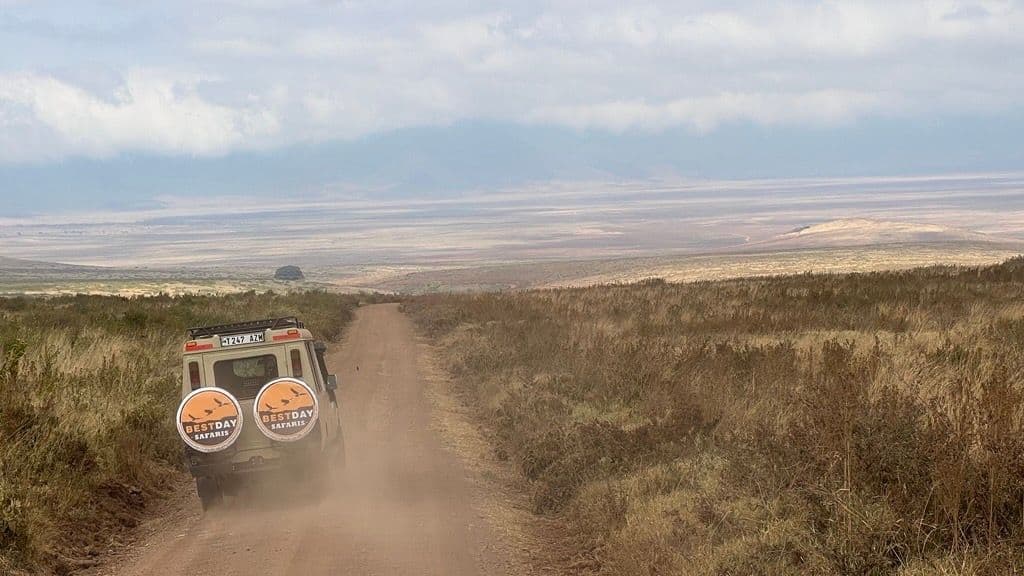 Una jeep da safari percorre una strada sterrata e polverosa attraverso una vasta savana, con montagne lontane sotto un cielo nuvoloso.