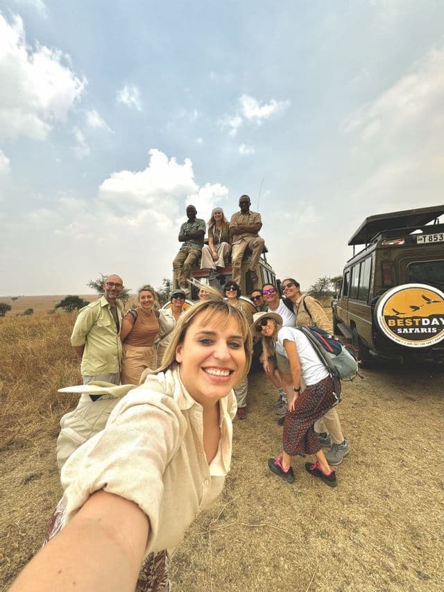 Un gruppo WeRoad in viaggio sorride per un selfie, radunato su e intorno a una jeep da safari in una savana erbosa.
