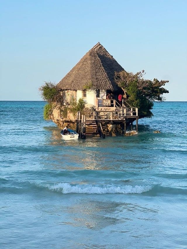 Un edificio con tetto di paglia su una formazione rocciosa nel mare, con una piccola barca ormeggiata alla sua scala in legno.