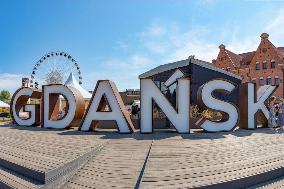 Ein großes, dreidimensionales 'GDAŃSK'-Schild auf einem Holzdeck, mit Riesenrad und Gebäuden im Hintergrund.