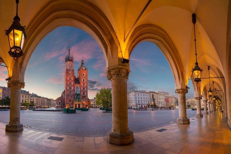 Eine historische Backsteinkirche mit zwei Türmen steht auf einem Stadtplatz, bei Sonnenuntergang von unter beleuchteten Steinbögen aus gesehen.