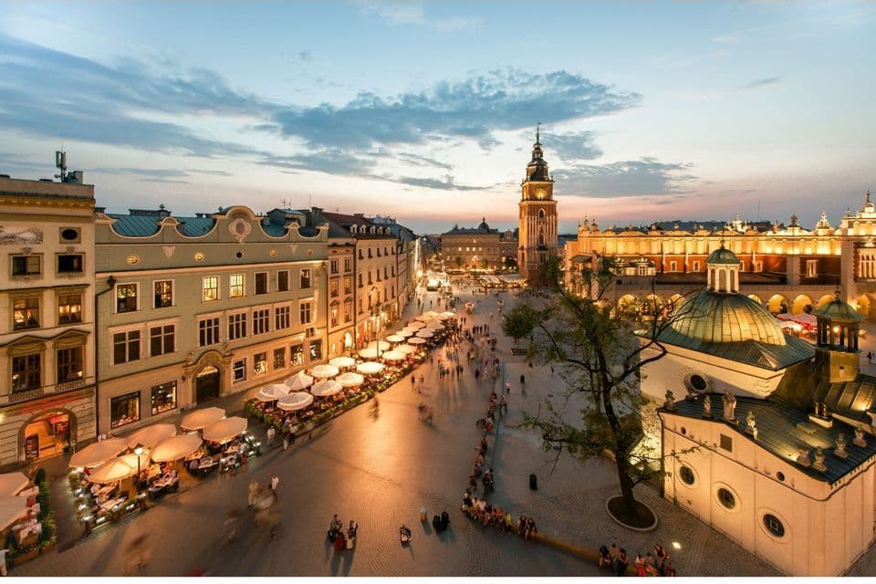 Ein erhöhter Blick auf einen historischen europäischen Stadtplatz in der Dämmerung, mit beleuchteten Gebäuden, Straßencafés und flanierenden Menschen.