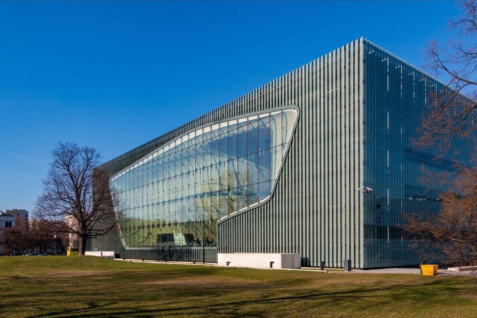 Ein großes, modernes Gebäude mit einer einzigartigen Glasfassade und vertikal gerippten Paneelen, auf einem grünen Rasen unter einem klaren blauen Himmel gelegen.