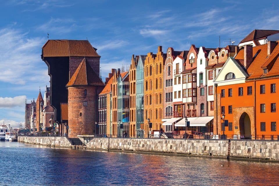 Bunte historische Gebäude und ein großer Backstein-Hafenkran säumen an einem sonnigen Tag eine Flussuferpromenade.