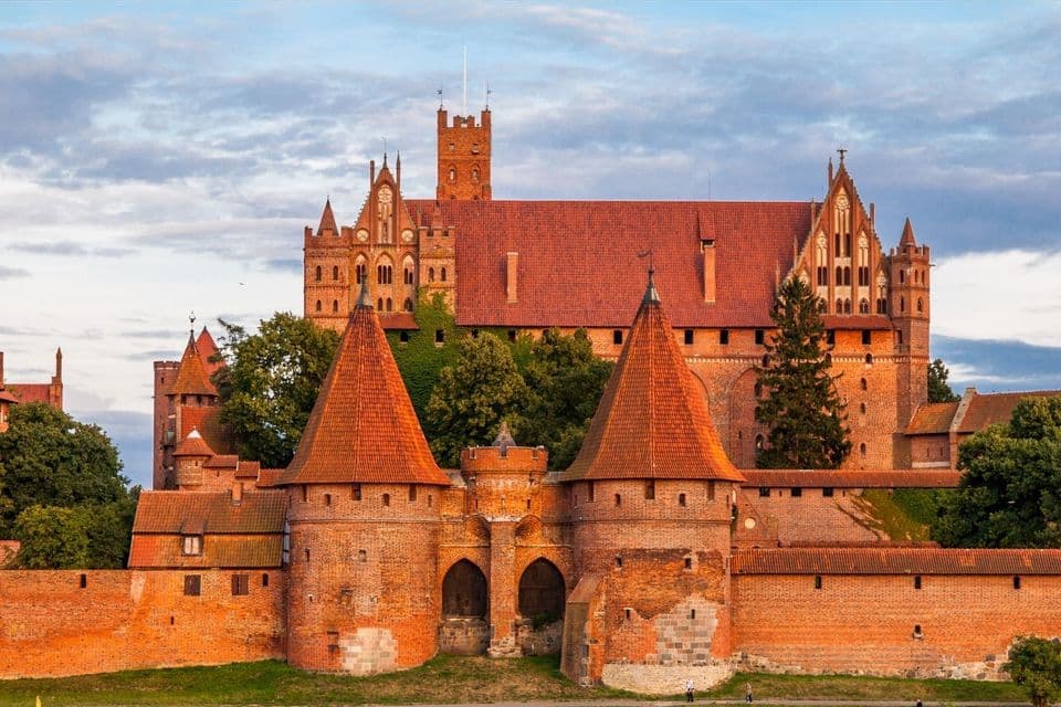 Ein großes mittelalterliches Backsteinschloss mit zahlreichen Türmen und einem befestigten Torhaus steht unter einem teils bewölkten Himmel.