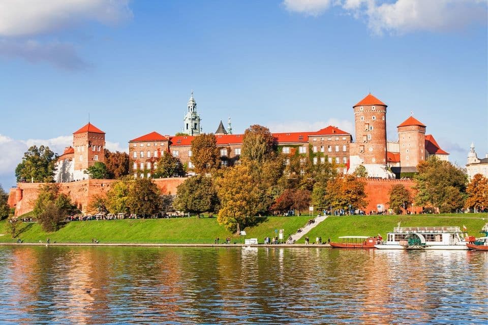 Ein historisches rotes Backsteinschloss mit Türmen steht an einem grasbewachsenen Flussufer, umgeben von Herbstbäumen unter blauem Himmel.