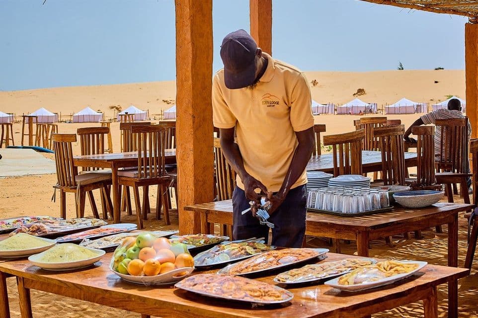 Un hombre con polo amarillo prepara un gran bufé al aire libre en mesas de madera en un campamento en el desierto con dunas de arena de fondo.